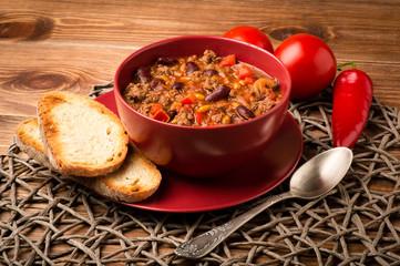 Chili con carne served in the red bowl on the wooden background.