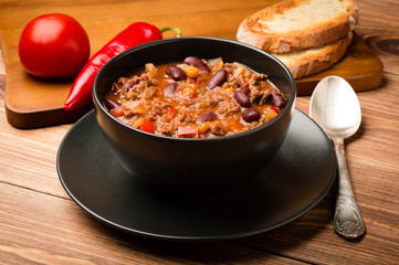 Chili con carne served in the black bowl on the wooden background.