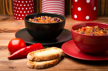 Chili con carne served in the red and black bowl on the wooden background.