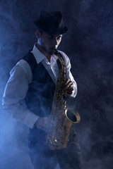Naklejka na ściany i meble Elegant saxophonist plays jazz on dark background in blue smoke