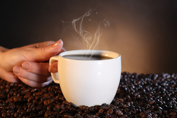 Woman holding cup of hot coffee among coffee beans on dark background