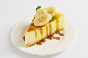 Slice of cheesecake topped with caramel sauce, banana and mint on plate on white table, close up