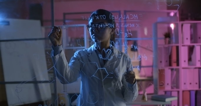Girl in lab coat writing formula in dark chemistry laboratory 