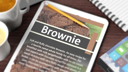Tabletop with various objects focused on tablet with recipe of "Brownie" on screen