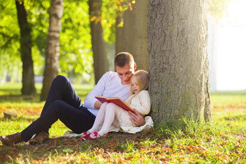 Fototapeta premium Cute little blonde girl sit near tree with father, read favorite