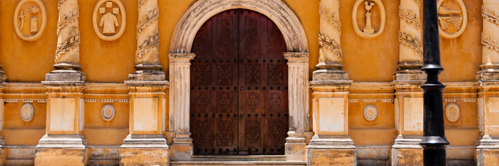 Old latern in front of yellow church with big brown wooden door, stone scarving and glass window
