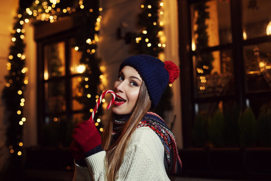 Night street portrait of a smiling  beautiful young woman biting Christmas candy cane and looking at camera. Lady wearing classic winter knitted clothes. Festive Christmas garland lights. Close up.