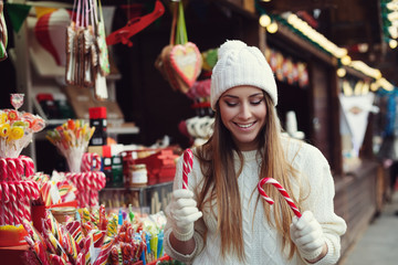 Street portrait of smiling beautiful young woman choosing candy canes on the festive Christmas fair. Lady wearing classic stylish winter knitted clothes. Close up