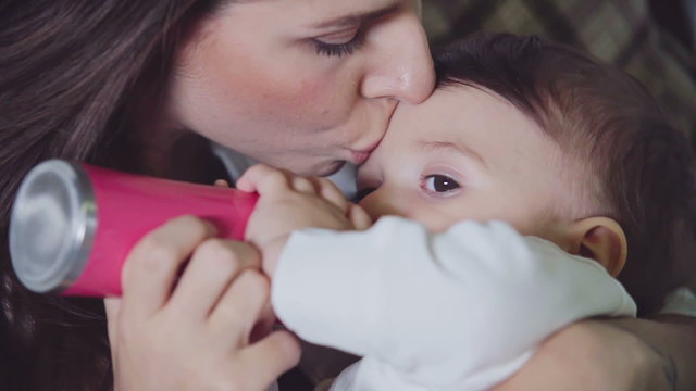 Young mother smooching forehead of her little son while he is drinking milk from baby bottle