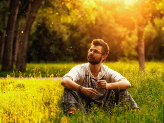 Naklejka na ściany i meble A bearded man sitting on a grass in the park on summer day, looks lost in thought and inspired