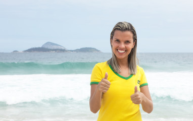 Blonde Frau im Brasilien-Trikot am Meer zeigt beide Daumen