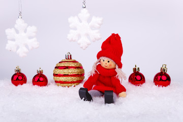 Xmas or new year composition with holiday decorations - little toy man sitting on the snow and little cristmas red baubles with snowflakes on the background. Christmas card