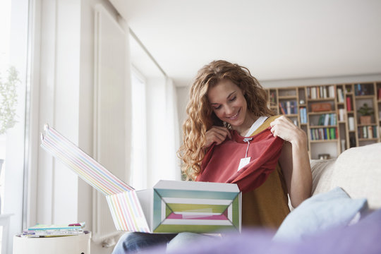 Smiling woman at home sitting on couch unpacking parcel with garment