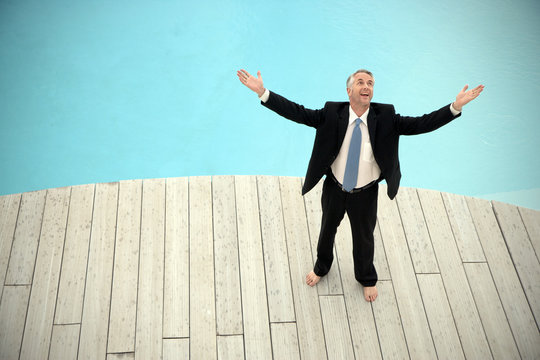 Barefoot businessman wearing black suit standing in front of swimming pool with arms raised - Powered by Adobe