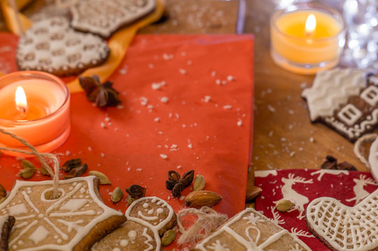 Christmas ginger cookies covered with white icing on a light brown wooden background and red stand with spices all over . They are illuminated by white lights and orange candles.