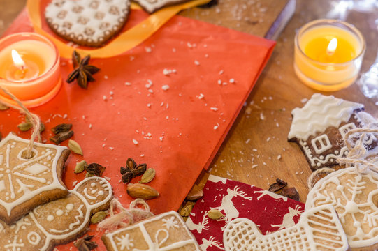 Christmas ginger cookies covered with white icing on a light brown wooden background and red stand with spices all over . They are illuminated by white lights and orange candles. 