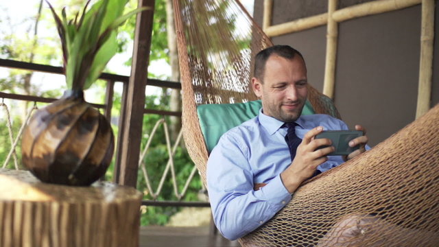 Happy businessman playing game on smartphone on hammock on terrace
