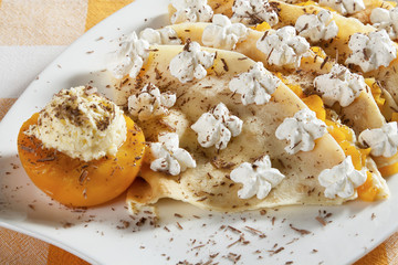 Crepes stuffed with vanilla cream and peach, decorated with whipped cream and grated chocolate, served on a white plate.