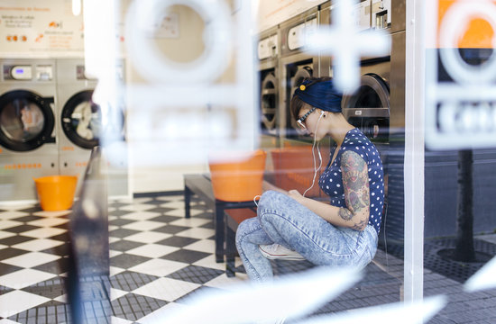 Tattooed young woman hearing music with earphones in a launderette