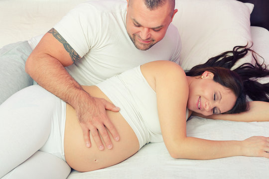 Pregnant smiling woman and husband lying in bed touching belly