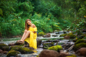 Девушка в желтом платье сидит в лесу на камне возле горного ручья