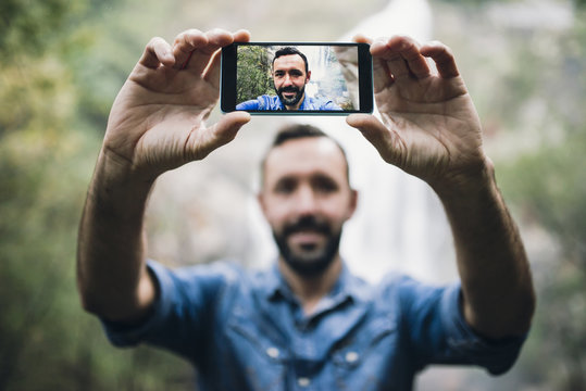 Bearded man taking a selfie with smartphone in front of a waterfall