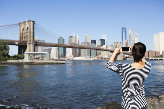 USA, New York City, back view of young woman taking a photo of Brooklyn Bridge with smartphone