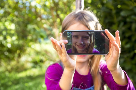 Little girl sitting on a meadow in the garden showing display of smartphone with her selfie