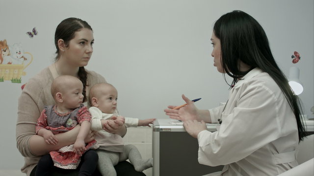 Female pediatrician doctor explain something to mother with newborn baby twins at modern hospital indoors