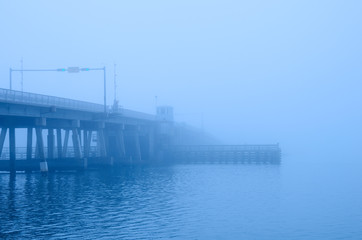 A draw bridge with stop light, gate, bridge keeper's house and channel bumpers is disappearing into a heavy fog.