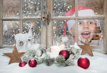 Kind schaut freudig überrascht aus weihnachtlich dekoriertem Winterfenster, innen leuchtet schon...