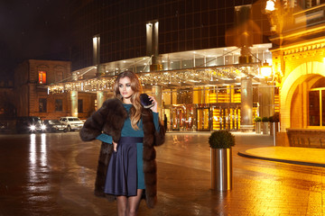 Beautiful young sexy blonde wearing evening makeup in elegant fitting dress fashionable stylish expensive fur coat walk night street lights evening hand  handbag luxury rich life going party concert