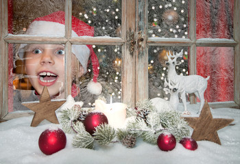 Kind schaut freudig überrascht aus weihnachtlich dekoriertem Winterfenster, innen leuchtet der...