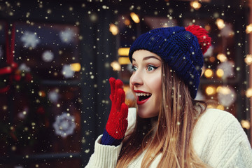 Street emotional portrait of young beautiful woman looking surprised. Model looking aside. Lady wearing stylish classic winter knitted clothes.  Festive garland lights. Magic snowfall effect. Close up