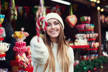 Young beautiful smiling woman holding Christamas candy cane. Festive fair as background. Selective focus on the model. The object is defocused. Close up
