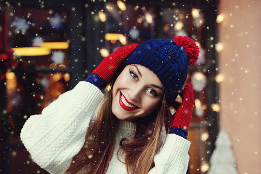 Street portrait of smiling beautiful young woman wearing classic winter knitted clothes. Model looking at camera, Festive  garland lights. Magic snowfall effect. Close up. Toned