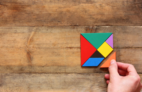 Top View Photo Of Man's Hand Holding A Missing Piece In A Square Tangram Puzzle, Over Wooden Table.
