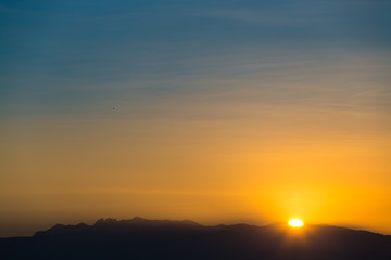 Fototapeta na wymiar Sun behind dark mountain silhouettes, with blue and orange colored sky