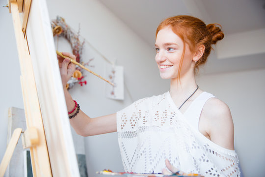 Smiling attractive woman painter with red hair painting on canvas