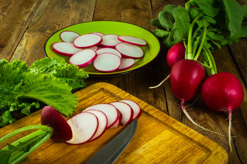 A bunch of fresh radishes, lettuce leaves, slices of radish and knife on old chopping board