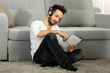 Young man listens music with headphones on the floor against grey sofa in the room
