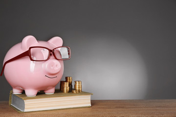 Piggy bank in glasses with book and coins on grey background
