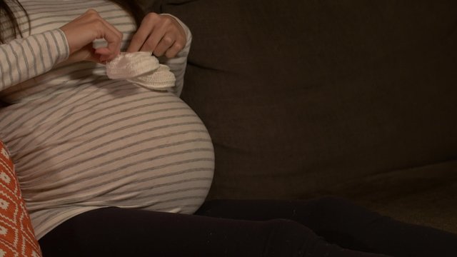 Pregnant woman on couch playing with baby shoes on tummy- pan shot