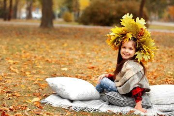 Happy young girl in yellow autumn wreath sitting on plaid in park
