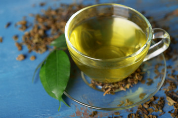 Glass cup of tea with green leaves and scattered tea around on blue wooden background