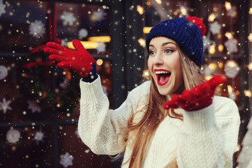 Street portrait of young beautiful woman acting thrilled, wearing stylish knitted clothes. Model expressing joy and excitement with hands and face. Festive garland lights. Snowfall effect. Close up.