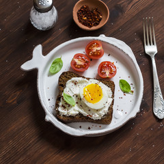 Sandwich with feta cheese and boiled egg, tomatoes, and basil on a white plate on a dark wooden surface. Healthy breakfast or snack