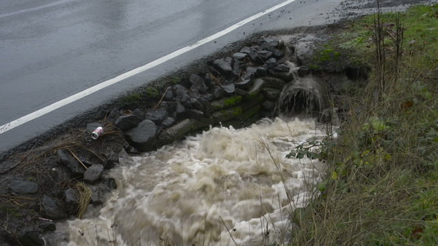 Water surging out of a pipe into a roadside drainage ditch, car passes by on the roadway