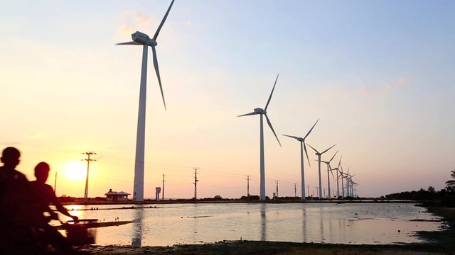 
Sunset view of salt ponds and turbines from wind farm with tuk tuk with people on bicycles passing in foreground 