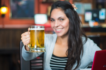 Brunette model sitting by restaurant table holding glass of beer and posing with positive attitude smiling